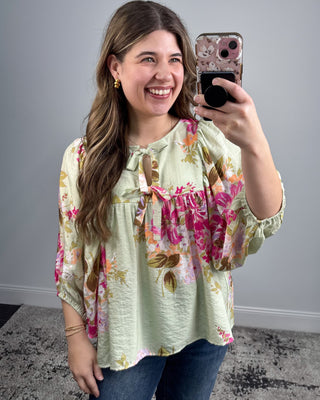 Woman taking a selfie wearing a floral blouse against a gray wall.