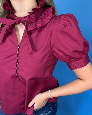 A close-up of a person wearing a deep magenta blouse with a ruffled collar, tie detail at the neckline, puff sleeves, and decorative buttons down the front, paired with blue jean