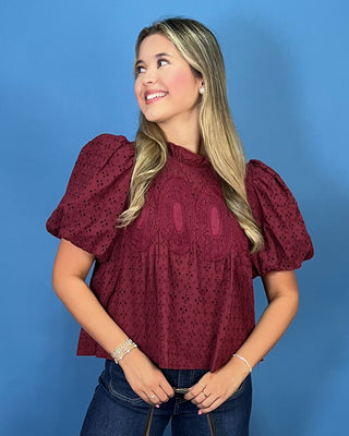 A woman with blonde hair wearing a burgundy eyelet puff-sleeve top and blue jeans