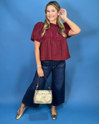 A woman modeling a short-sleeve burgundy eyelet lace top, dark blue wide-leg jeans, and metallic silver flats, holding a small metallic gold handbag
