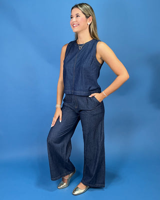 A woman modeling a dark blue denim two-piece set consisting of a sleeveless top and matching wide-leg pants, standing against a solid blue studio backdrop.