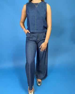 A woman modeling a dark blue denim sleeveless set with wide-leg pants, standing against a solid blue studio backdrop.