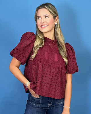 A woman modeling a burgundy eyelet puff-sleeve blouse with a high mock neck, paired with blue jeans