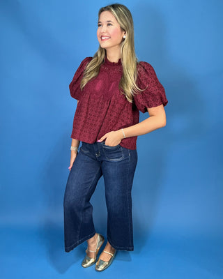 A woman modeling a burgundy eyelet puff-sleeve blouse with dark blue wide-leg jeans and metallic gold flats, 