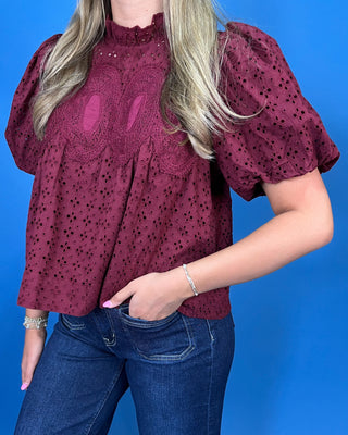 A woman wearing a burgundy eyelet blouse with puff sleeves and a ruffled high neck, paired with blue jeans