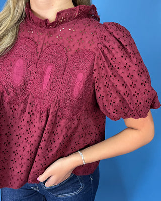 A close-up of a woman wearing a dusty rose eyelet blouse with puff sleeves and a ruffled high necklin