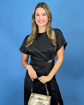 A woman posing in a black short-sleeve satin dress with a gold belt buckle, holding a metallic gold handbag against a solid blue background.