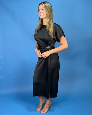 A woman modeling a black midi dress with short sleeves and a matching belt featuring a decorative gold-tone buckle, posed against a solid blue studio backdrop.