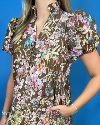 A woman wearing a brown short-sleeve blouse with a colorful floral and botanical print, featuring puffed short sleeves and a ruffled collar