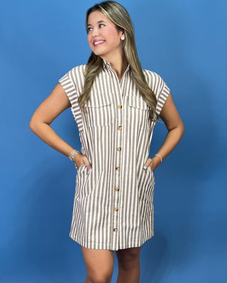 A woman modeling a sleeveless button-front shirt dress with vertical beige and white stripes, standing against a solid blue studio background.