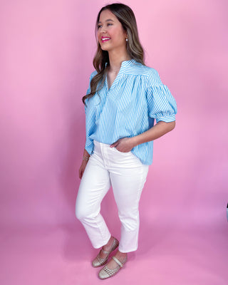 Woman wearing a blue striped blouse and white pants against a pink background