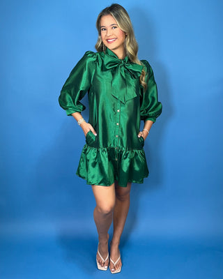 Emerald green satin mini dress with bow-tie neckline, puff sleeves, button front, pockets, and ruffle hem.