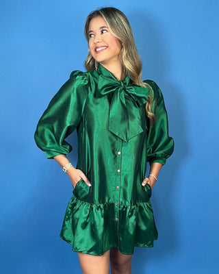 Emerald green satin mini dress with bow-tie neckline, puff sleeves, button front, pockets, and ruffle hem.
