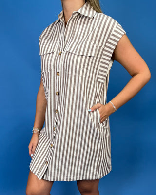 A woman wearing a sleeveless, button-front shirt dress with vertical brown and beige stripes, standing against a solid blue background.