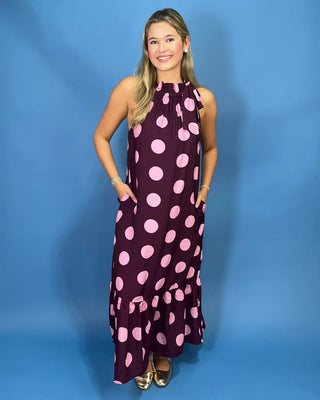 A woman modeling a sleeveless halter-neck maxi dress with a dark purple base and large light pink polka dots, standing against a solid blue studio backdrop.