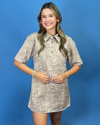 A young woman modeling a short-sleeve button-up shirt dress with a beige and white speckled pattern, standing against a solid blue studio background.