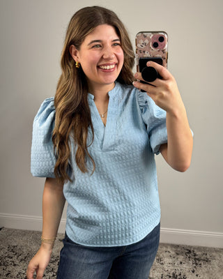 Woman taking a mirror selfie wearing a light blue textured top and jeans.
