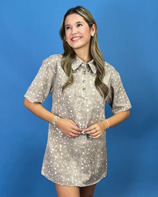 A woman modeling a short-sleeve button-up shirt dress with a beige and white speckled pattern, standing against a solid blue studio background.