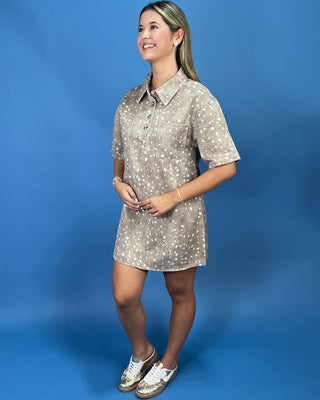 A woman modeling a short-sleeve collared shirt dress with a beige and white speckled pattern, standing against a solid blue studio backdrop.