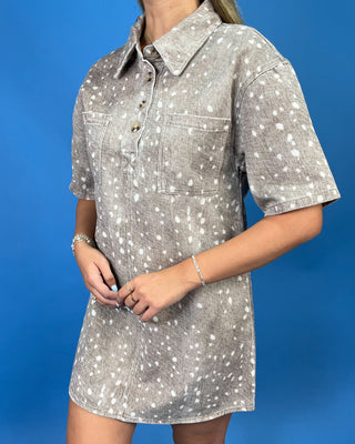 A woman wearing a short-sleeve collared shirt dress with a white speckled pattern on a gray base, standing against a solid blue background.