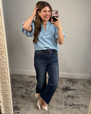 Woman taking a mirror selfie wearing a light blue top and dark blue jeans.