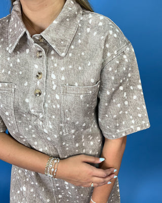 A close-up of a person wearing a short-sleeve collared shirt dress in a heather gray fabric with a white speckled or splattered pattern, featuring a button placket and chest pockets, set against a solid blue background.