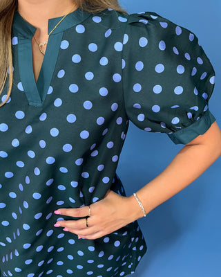 A close-up of a woman wearing a dark green blouse with light blue polka dots, featuring puff sleeves and a split V-neckline