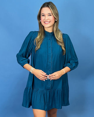 A woman modeling a teal blue long-sleeve mini dress with a button-up front and ruffled hem, standing against a solid blue studio background.