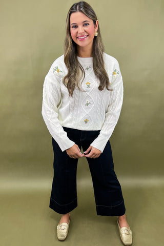 Woman wearing a white embroidered sweater and black pants against a green background