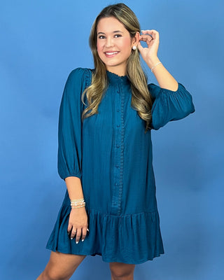 A woman modeling a teal blue long-sleeve mini dress with a ruffled collar, buttoned front, and ruffled hem, posed against a solid blue studio background.
