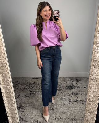 Woman taking a mirror selfie wearing a purple blouse and blue jeans.