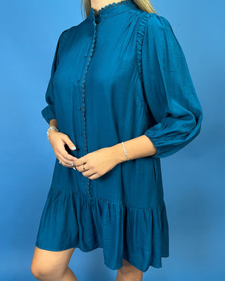 A woman wearing a teal blue long-sleeve mini dress with a scalloped ruffle collar, scalloped button placket, and ruffled hem, standing against a solid blue background.