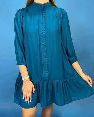 A woman wearing a teal blue long-sleeve mini dress with a scalloped collar, scalloped button placket, and a ruffled hem, shown against a solid blue background.