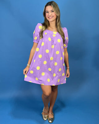A young woman modeling a lavender purple short-sleeve dress with yellow polka dots, standing against a solid blue studio backdrop.