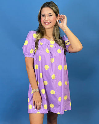 A woman modeling a lavender puff-sleeve mini dress with a yellow polka dot pattern, set against a solid blue studio background.