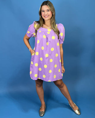 A woman modeling a lavender short-sleeve dress with yellow polka dots, standing against a solid blue studio backdrop.