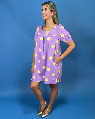 A woman modeling a lavender purple short-sleeve dress with yellow polka dots, standing against a solid blue studio background.