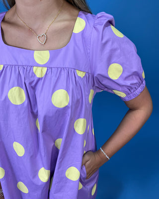 A close-up of a person wearing a lavender purple short-sleeve top with large light yellow polka dots, paired with a heart-shaped rhinestone necklace, against a solid blue background.