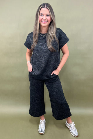 Woman wearing a dark gray t-shirt and wide-leg pants against a green background