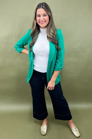 Woman wearing a green cardigan, white turtleneck, black pants, and white shoes on a green background