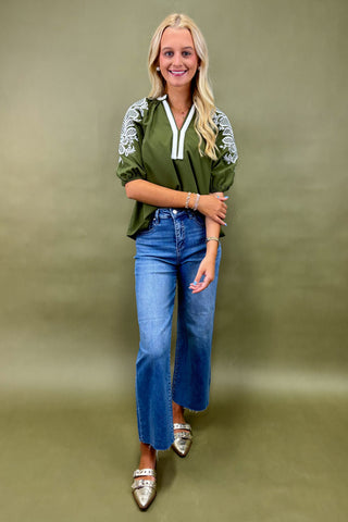 Woman wearing a green embroidered blouse and blue jeans against a green background
