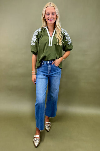 Woman wearing an olive green top with white embroidery and blue jeans on a beige background