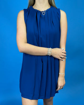 A woman wearing a sleeveless royal blue pleated mini dress with a gathered neckline and silver heart-shaped necklace, standing against a solid blue background.