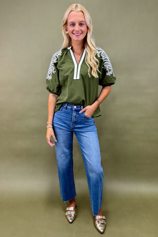 Woman wearing an olive green top with white embroidery and blue jeans on a beige background
