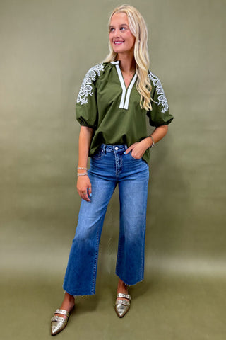 Woman wearing an olive green top with white embroidery and blue jeans on a beige background