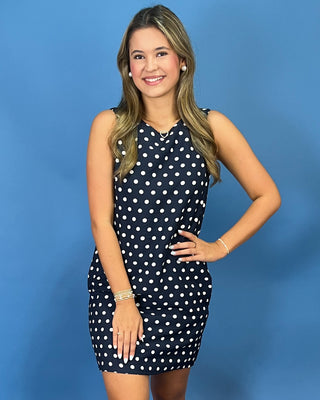 A woman modeling a sleeveless navy blue and white polka dot dress against a solid blue studio background.