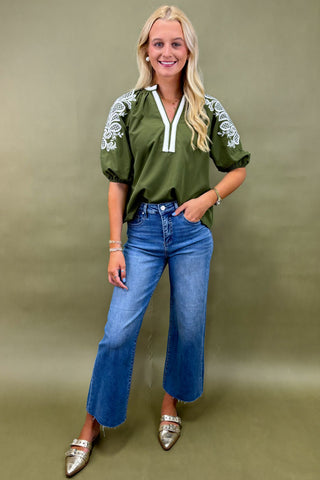 Woman wearing an olive green top with white embroidery and blue jeans on a beige background