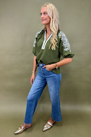 Woman wearing a green top with white embroidery and blue jeans on a green background