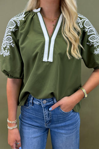 Woman wearing an olive green top with white embroidery and blue jeans on a beige background
