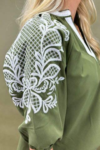 Woman wearing an olive green top with white embroidery and blue jeans on a beige background